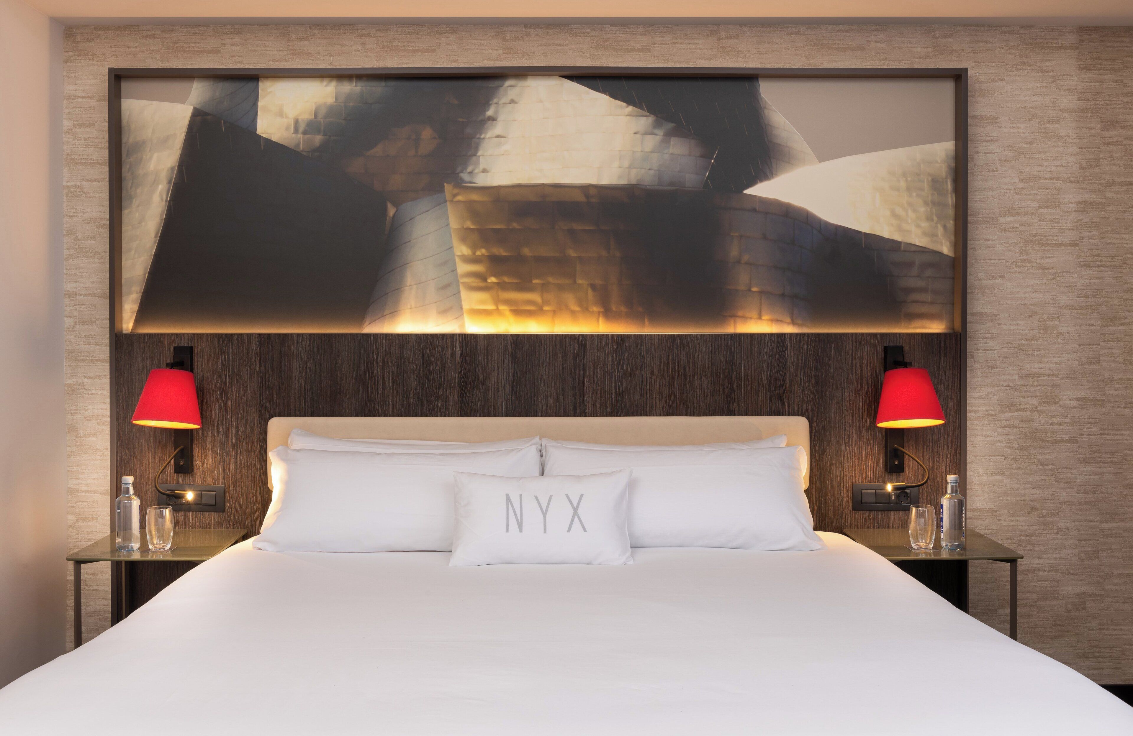 Ein Hotelzimmer mit einem großen Bett, zwei Nachttischen, einer roten Lampe auf jedem Tisch und einem Wandbild über dem Bett.