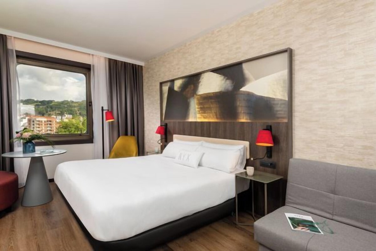 A hotel room with a large bed, a small table, a sofa, and a large window with curtains.