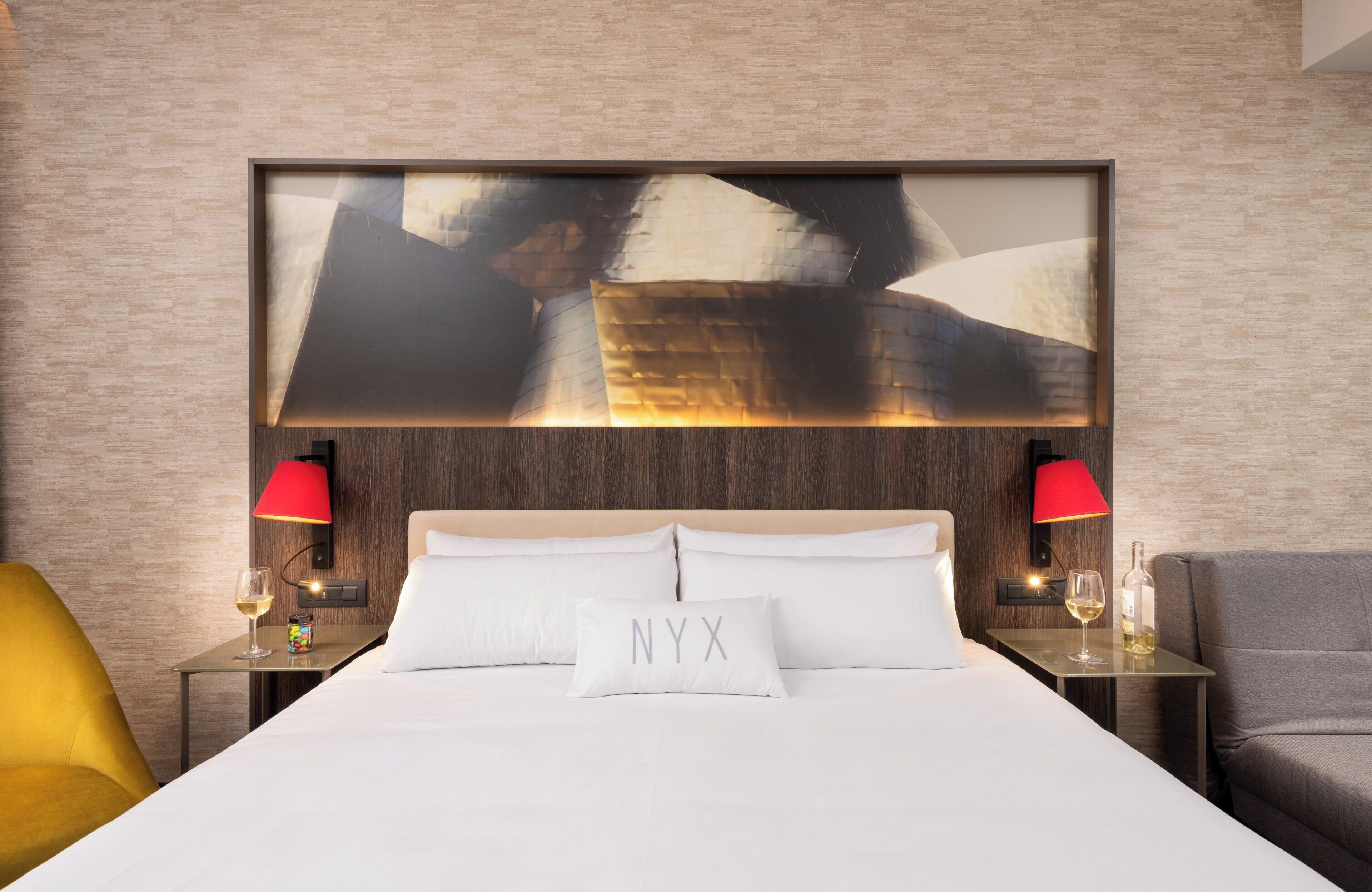 A hotel room with a large bed, two bedside tables with red lamps, a glass table with a wine glass, and a modern abstract painting above the bed.