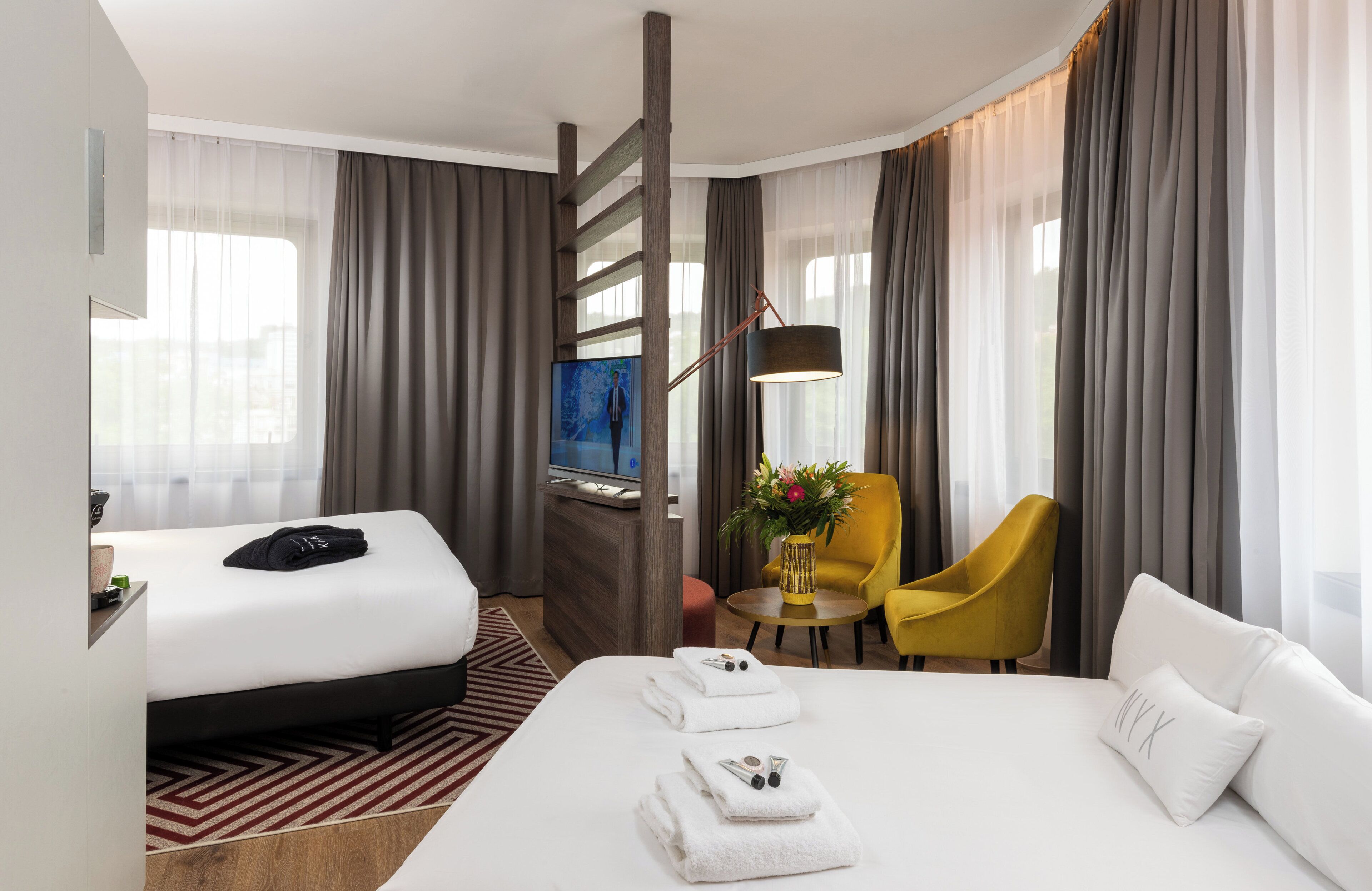 A hotel room with a large bed, a desk with a flat-screen TV, a yellow armchair, and a small table with a vase of flowers.