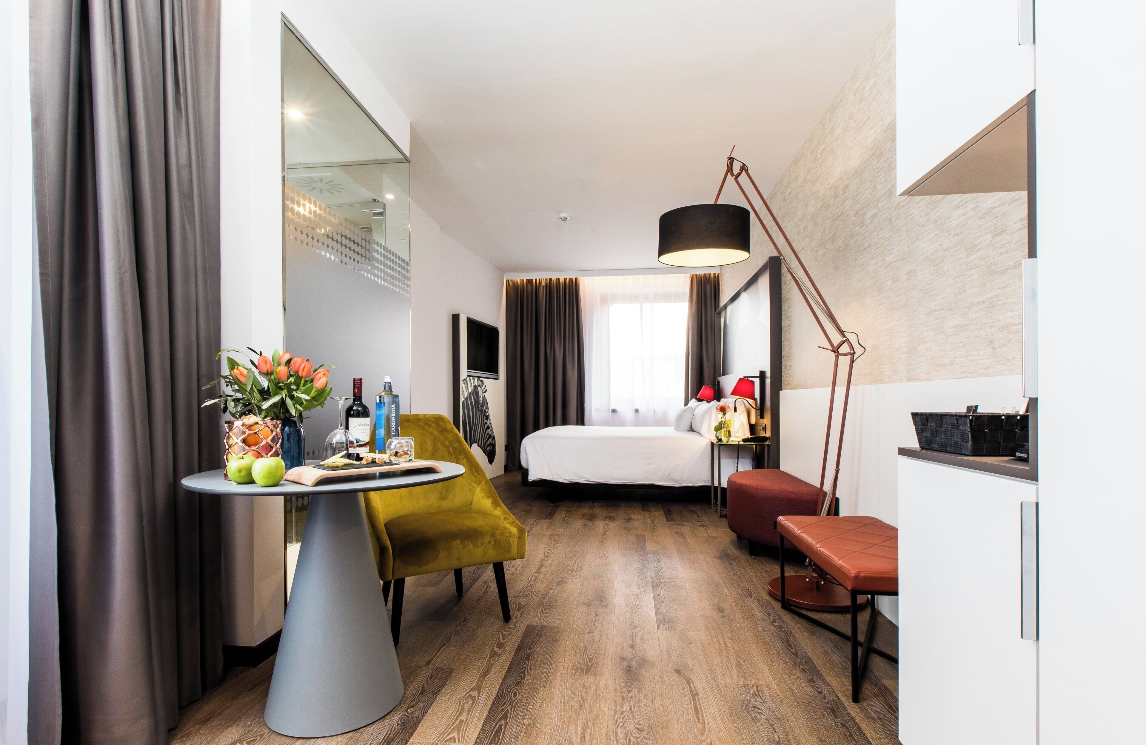 A modern hotel room with a bed, a desk, a chair, a table with apples and a vase, and a large mirror.