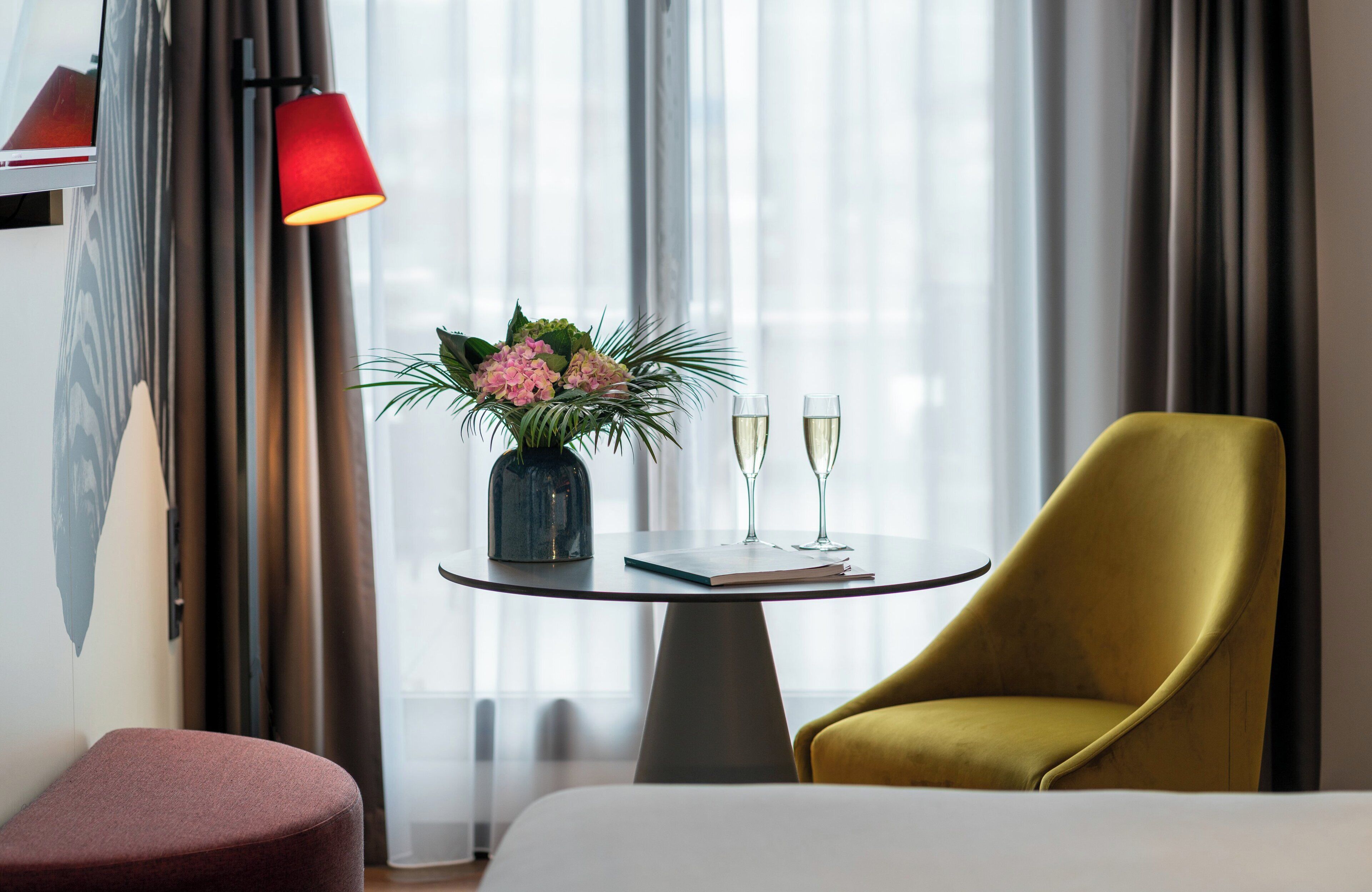 A hotel room with a bed, a round table with two glasses of champagne, a yellow armchair, a red lamp, and a vase with flowers.