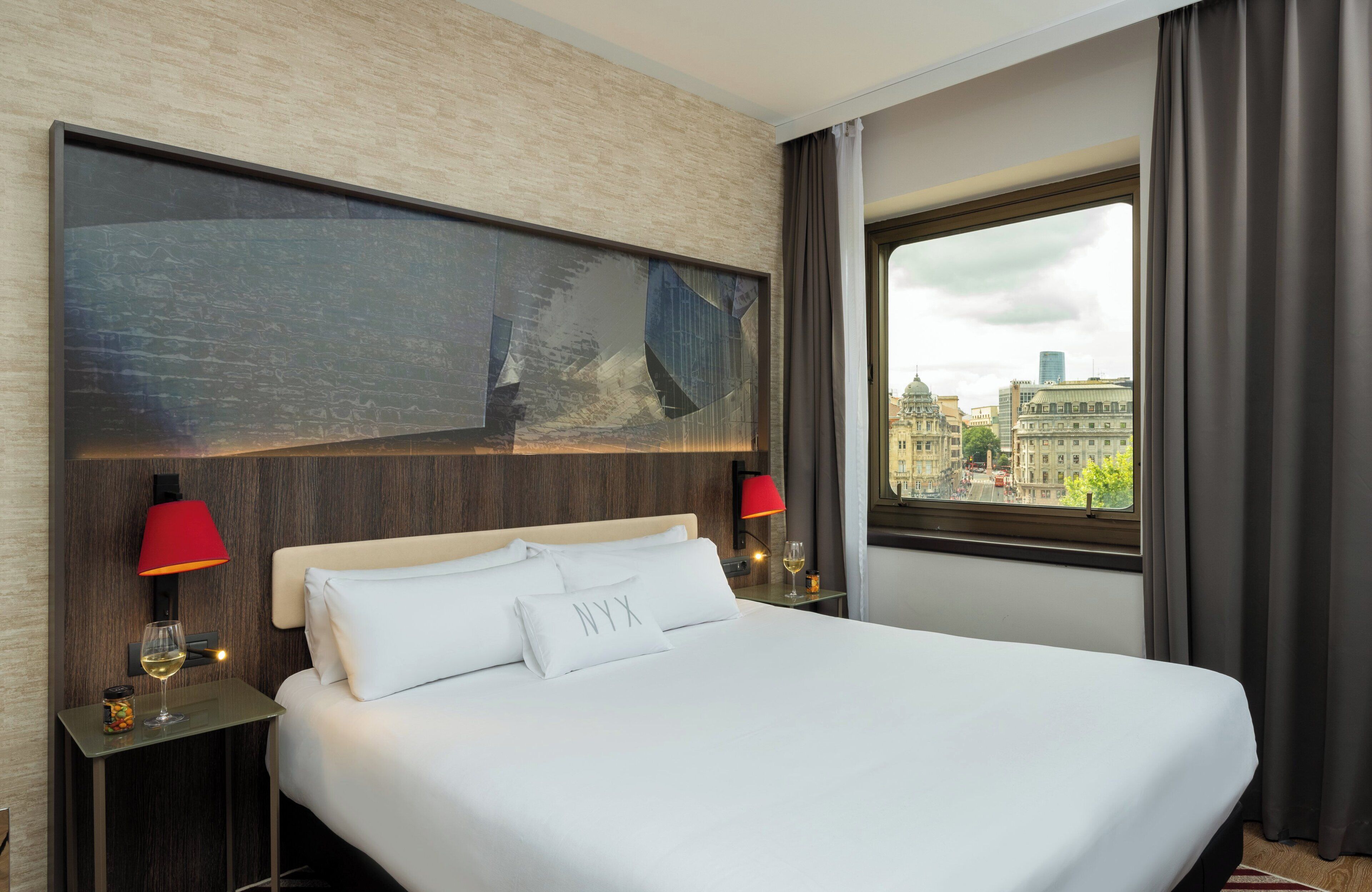 Habitación de hotel con una cama grande, dos mesitas de noche con lámparas, vista a la ciudad a través de una ventana y un cuadro abstracto de gran tamaño en la pared.