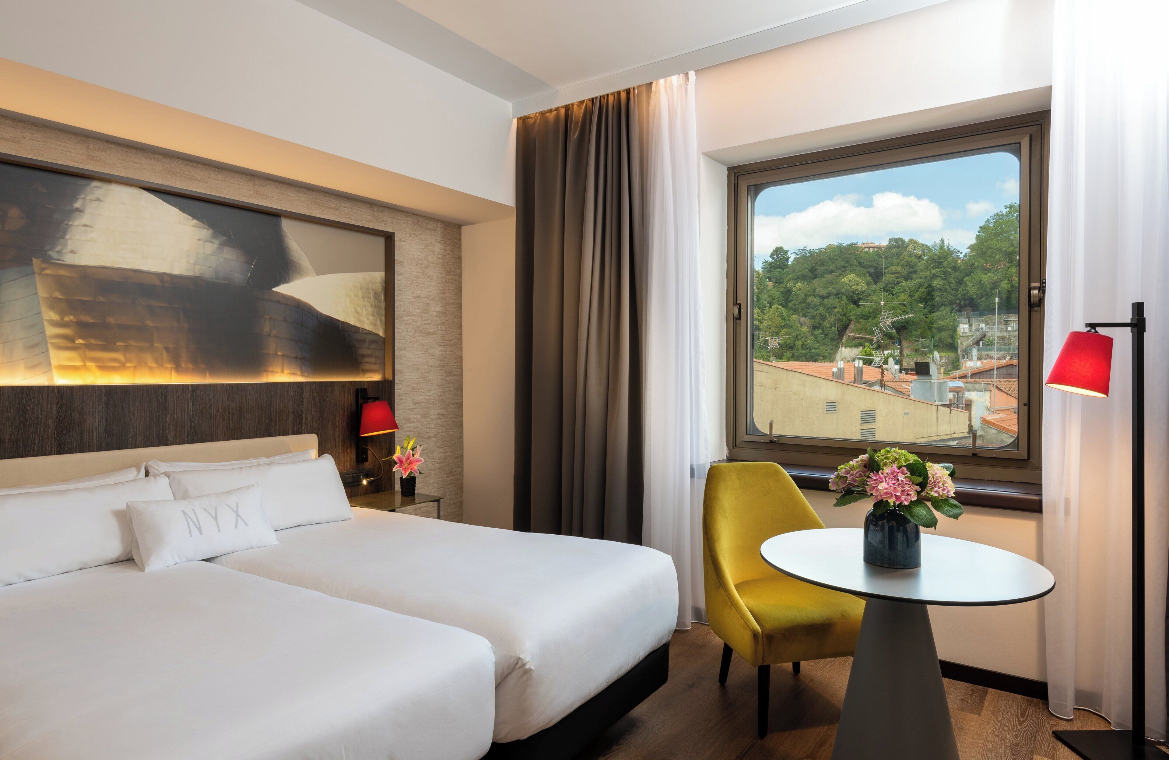 Une chambre d’hôtel avec un grand lit, une petite table ornée d’un vase de fleurs, une chaise jaune et une vue sur les bâtiments par la fenêtre.