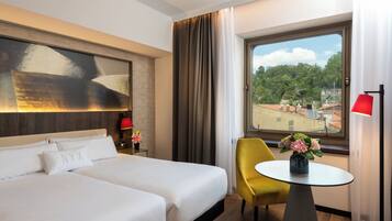 A hotel room with a large bed, a small table with a vase of flowers, a yellow chair, and a view of buildings through a window.