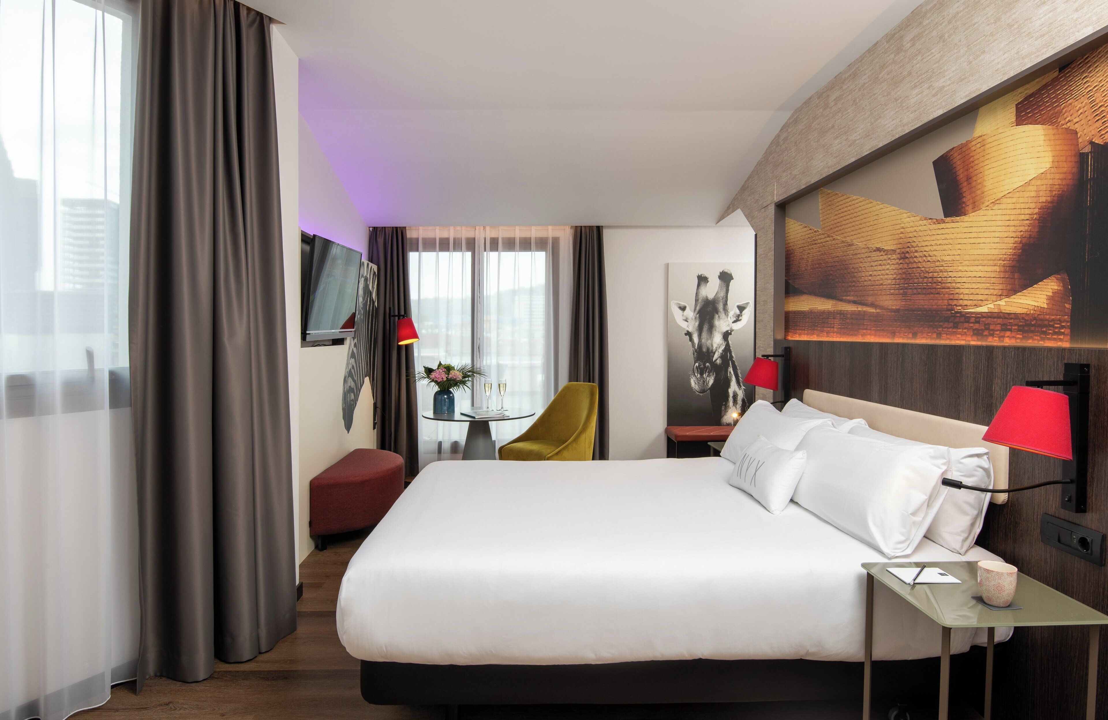 Habitación de hotel con una cama grande, un escritorio con lámpara, una silla y vistas a la ciudad.