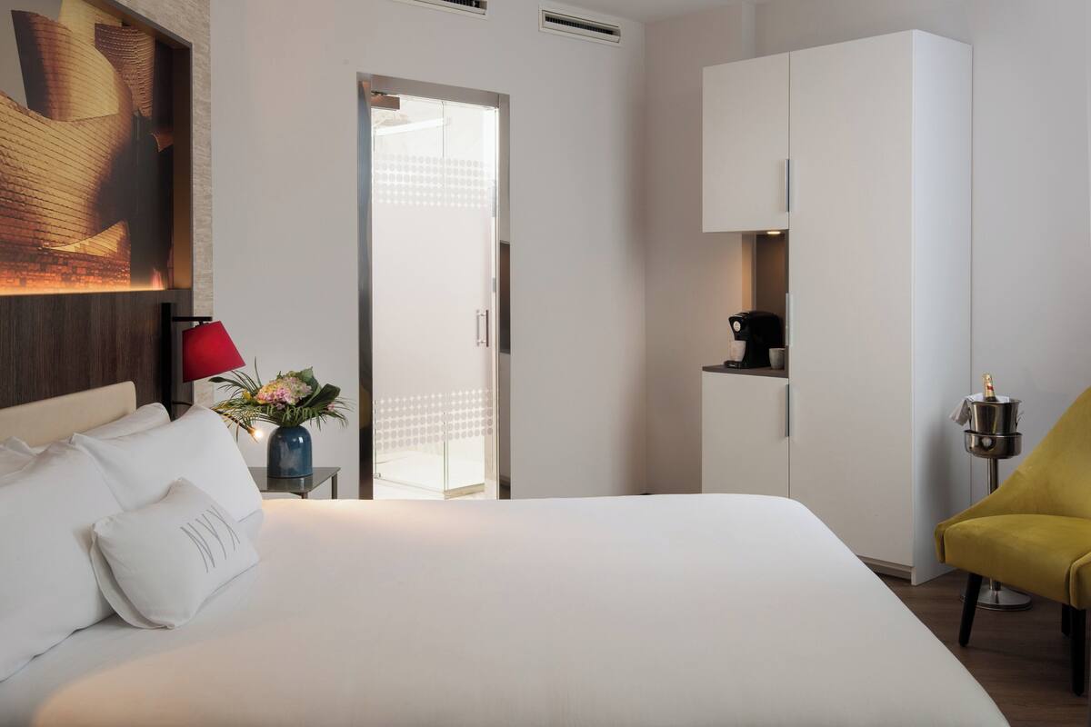 A hotel room with a bed, a red lamp, a yellow chair, a kitchenette, and a door leading to a balcony.