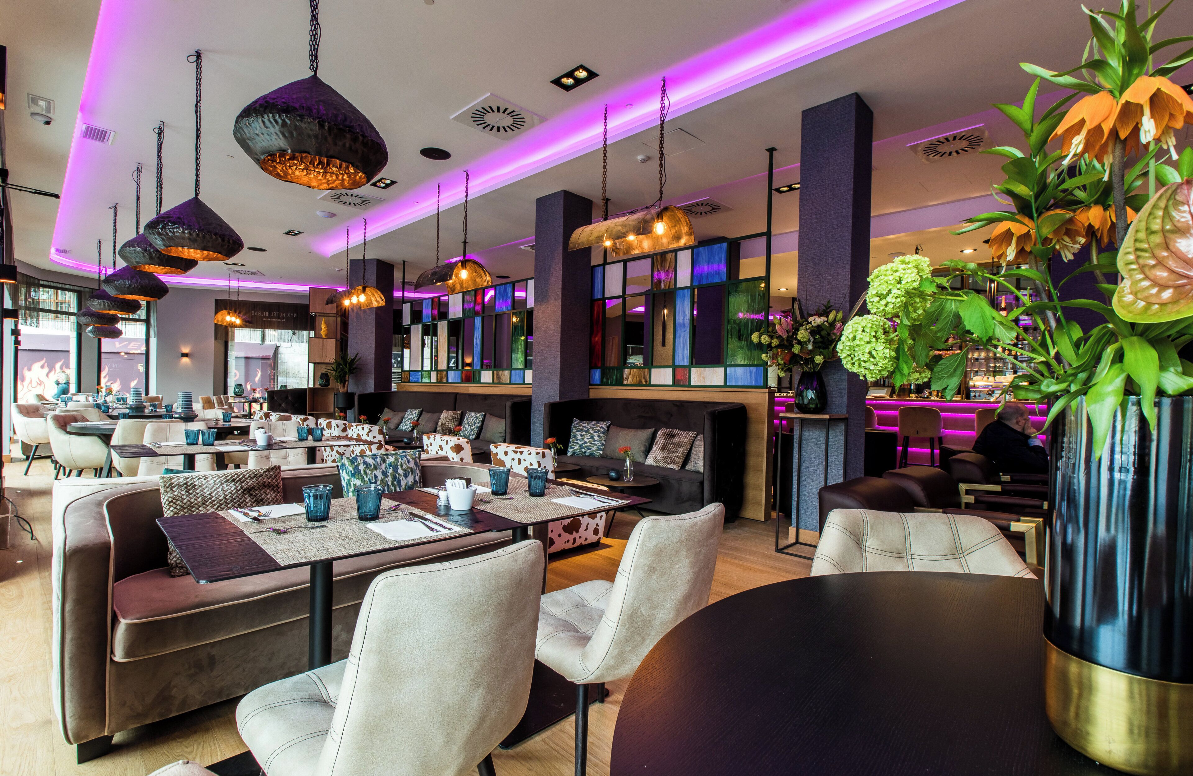 Un restaurante moderno con diversas opciones de asientos, incluyendo mesas y sofás, iluminación ambiental y plantas decorativas.