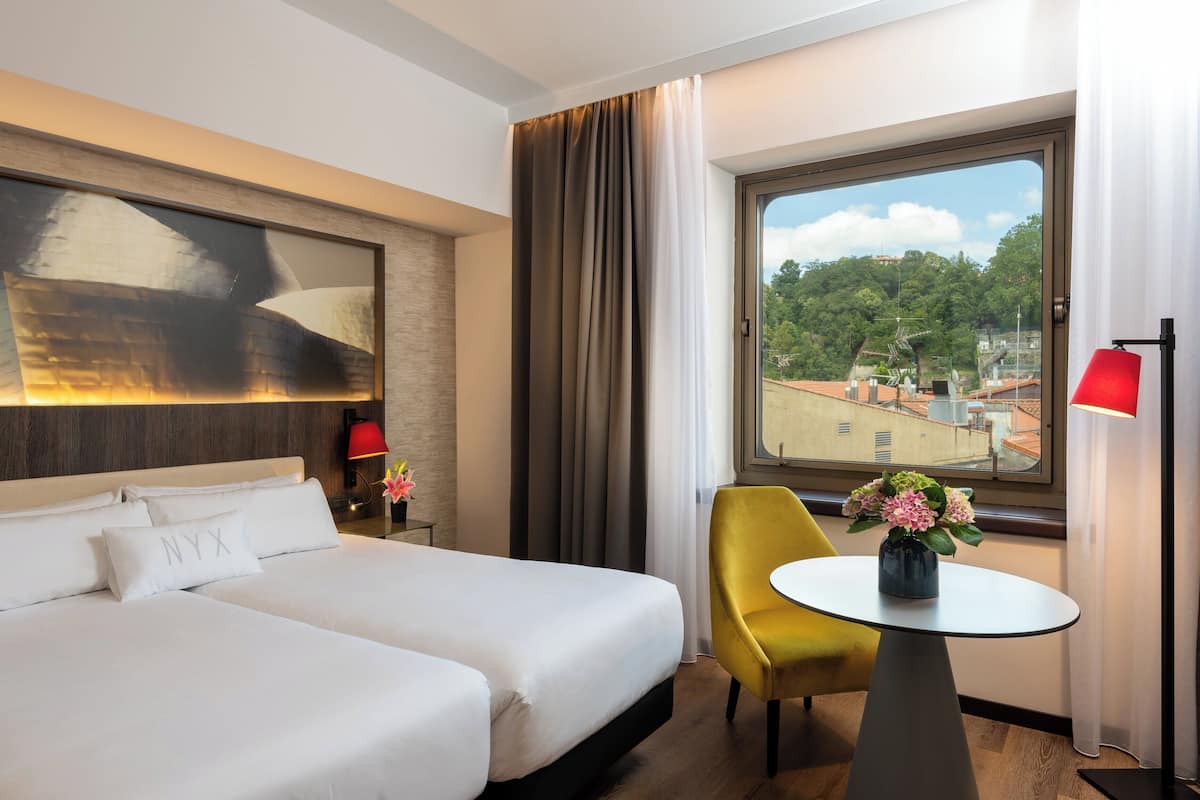 A hotel room with a large bed, a small table with a vase of flowers, a yellow chair, and a view of buildings through a window.