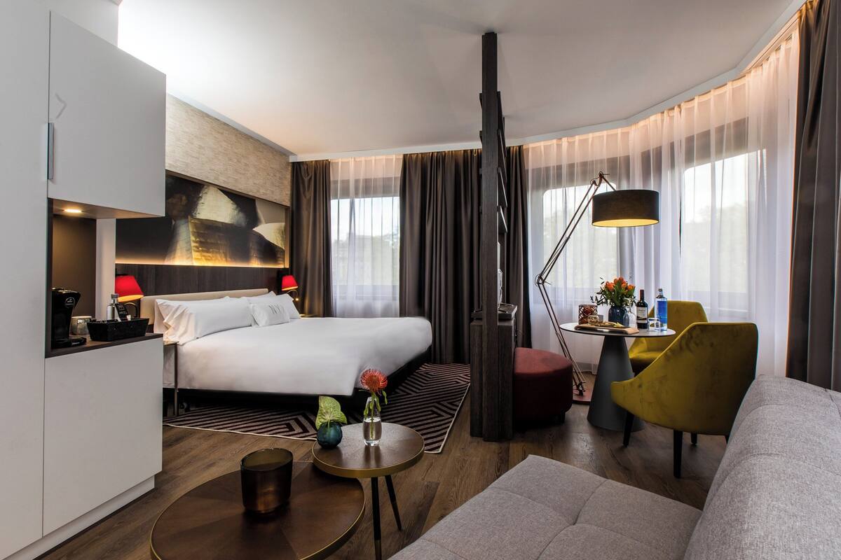 A modern hotel room with a bed, a desk, a sofa, and a dining area.