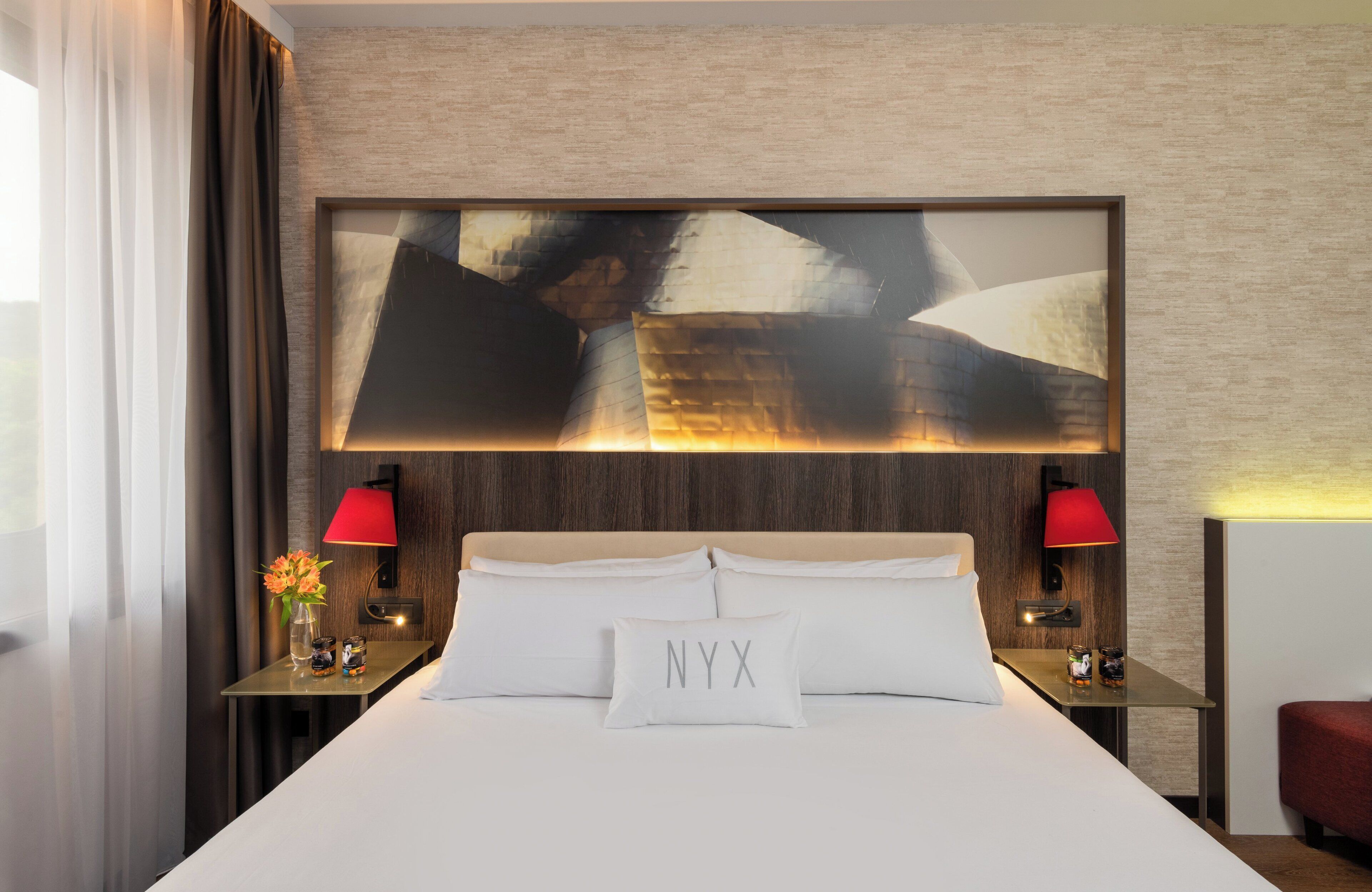 A hotel room with a large bed, two bedside tables, a red lamp, a wall-mounted artwork, and a nightstand with a vase and candles.