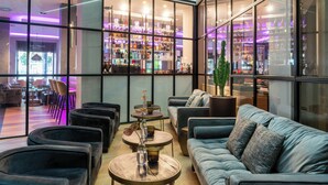 A modern lounge area with sofas, chairs, and tables, featuring a large window with a view of a bar and shelves stocked with bottles. - NYX Hotel Bilbao by Leonardo Hotels (Bilbao)