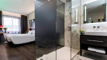 Une chambre d’hôtel moderne équipée d’un lit, d’une douche en verre, d’une coiffeuse avec un miroir et d’une petite plante.