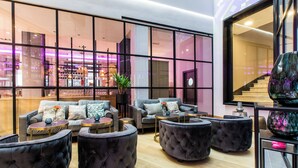 A modern lounge area with a sofa set, round ottomans, and a large window with a view of a bar area. - NYX Hotel Bilbao by Leonardo Hotels (Bilbao)