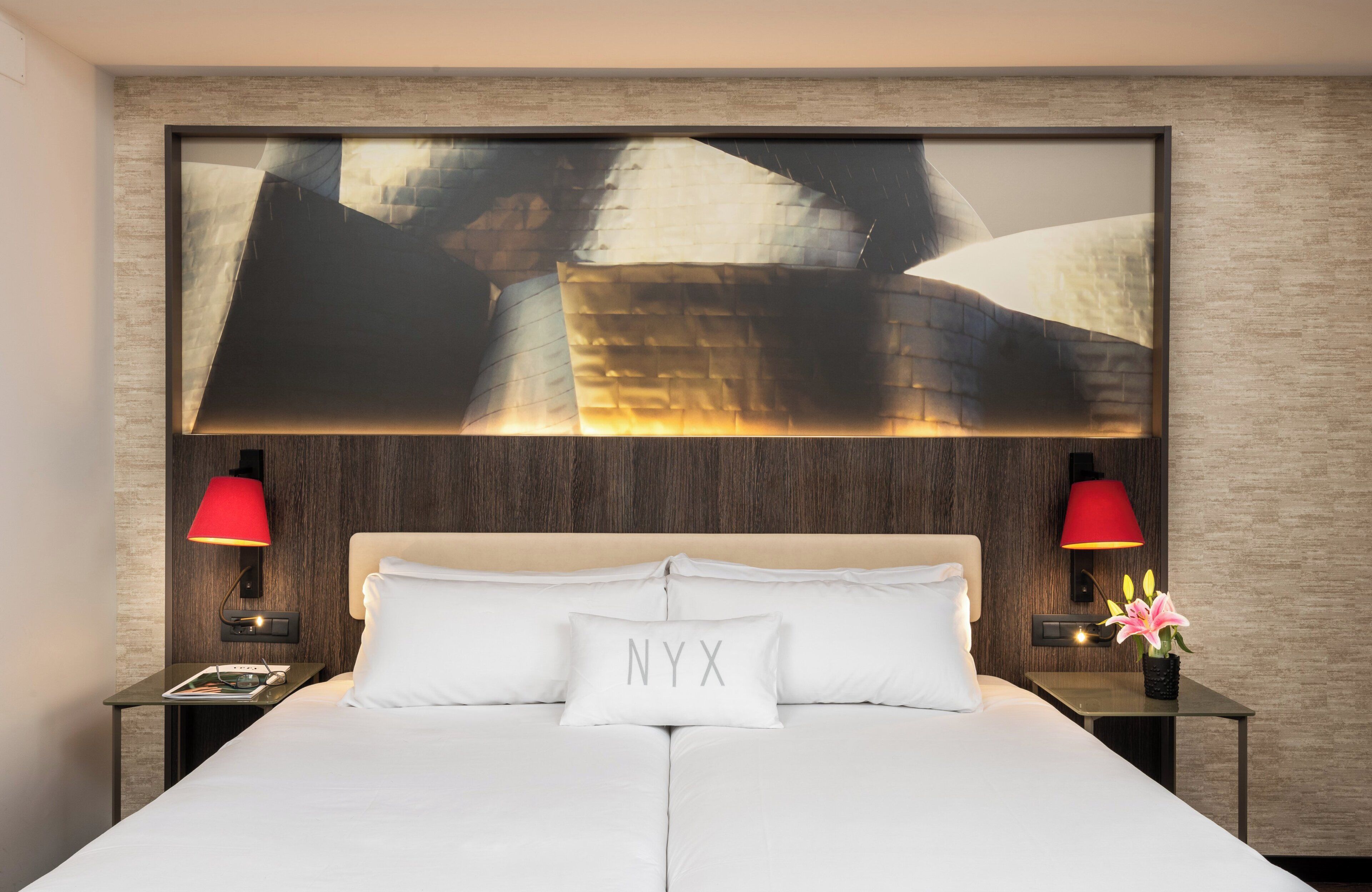 Une chambre d’hôtel avec un grand lit, deux lampes de chevet, une table de chevet avec un magazine et une œuvre d’art accrochée au mur.