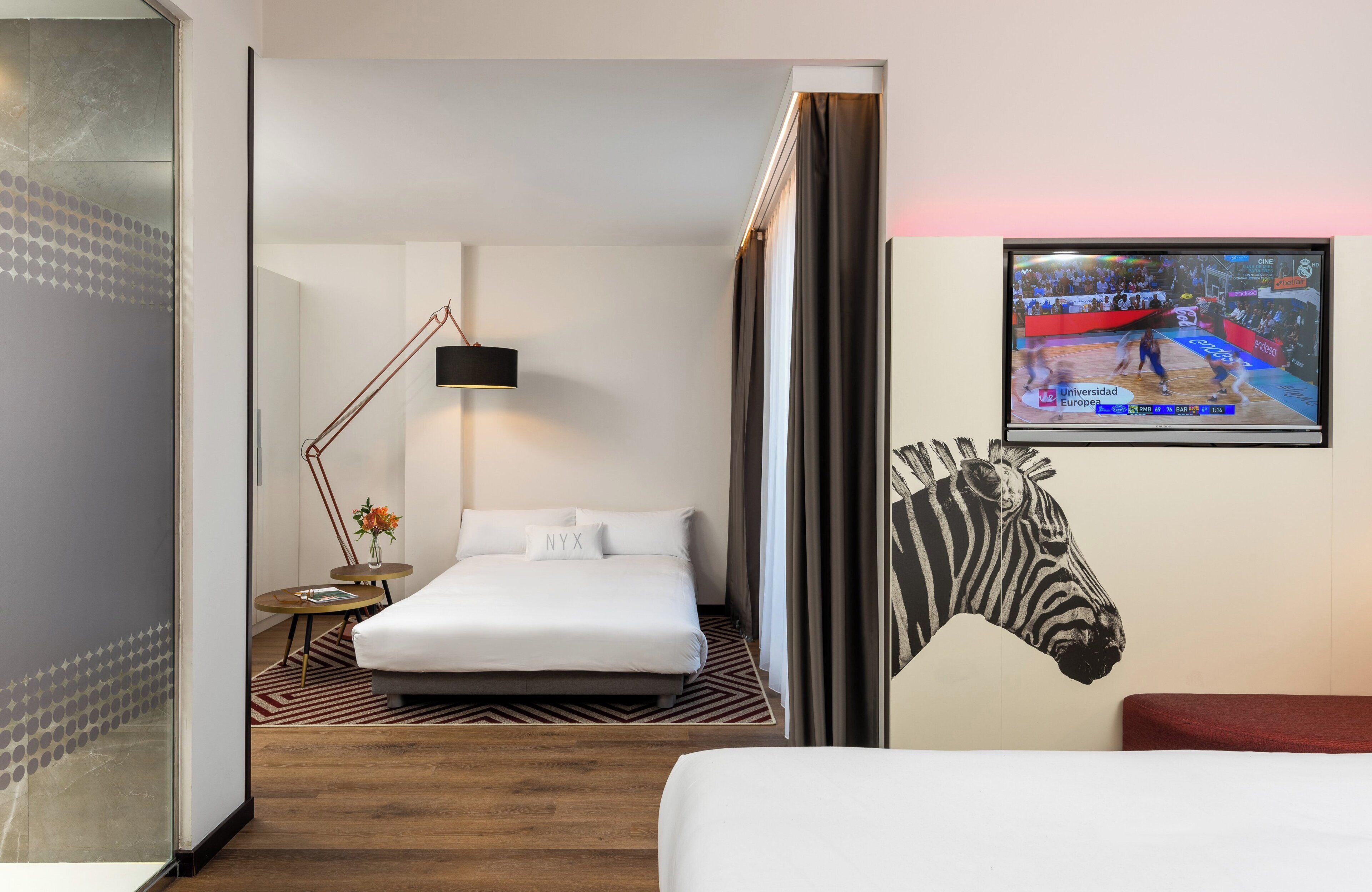 Habitación de hotel con cama, televisor mostrando un evento deportivo, una obra de arte con cebra y una lámpara moderna.