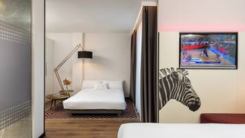 Habitación de hotel con cama, televisor mostrando un evento deportivo, una obra de arte con cebra y una lámpara moderna.