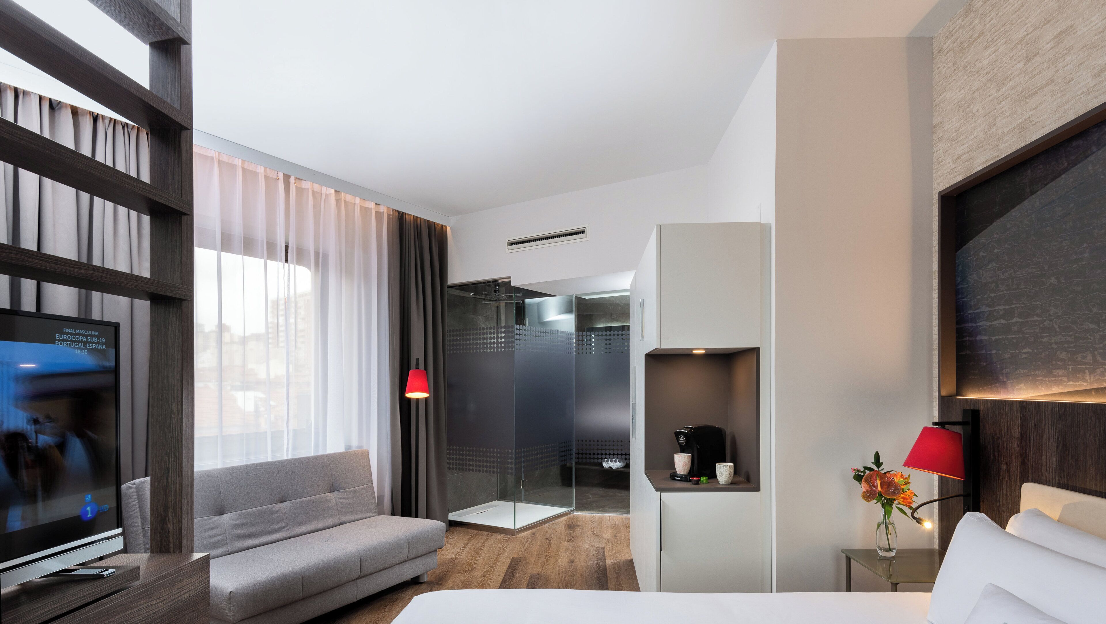 Habitación de hotel moderna con sofá gris, televisor, zona de ducha y cama.
