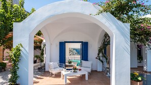 Ein Sitzbereich im Freien mit weißen Wänden, einem Tisch und Stühlen, umgeben von Blumentöpfen und Blick aufs Meer.