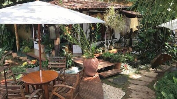 A patio area with wooden furniture, a table, and chairs under a white umbrella. There are potted plants and a small garden area with a stone pathway. - Pousada Bambu Brasil (Alto Paraíso de Goiás)