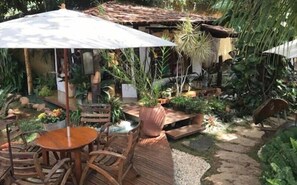 Ein Patio mit Holzmöbeln, einem Tisch und Stühlen unter einem weißen Sonnenschirm. Kübelpflanzen und ein kleiner Gartenbereich mit einem Kiesweg sind ebenfalls vorhanden.