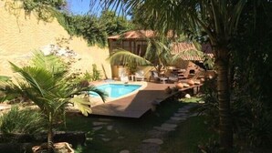 A pool area with a thatched umbrella, lounge chairs, and a small building with a tiled roof. - Pousada Bambu Brasil (Alto Paraíso de Goiás)