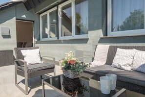 An outdoor seating area with wicker furniture and a glass table, a wicker basket with flowers, and a view of a house with large windows. - Miralior Apartment (Mainz)