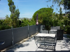 Um pátio externo com mesa e cadeiras de tampo de vidro, uma pérgula com guarda-sol, e vista para uma paisagem verde e colinas ao longe.