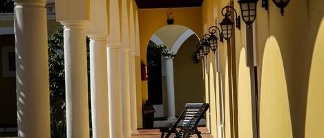 Un corredor cubierto con columnas blancas, bancos y faroles fijados a la pared.