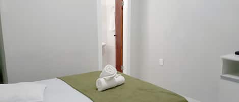 A hotel room with a bed, a green throw blanket, a white towel, a wooden door, and a white shelf.