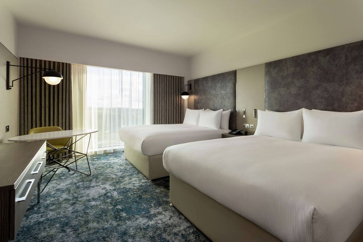 Quarto de hotel com duas camas, uma escrivaninha, uma cadeira e uma janela grande com cortinas.