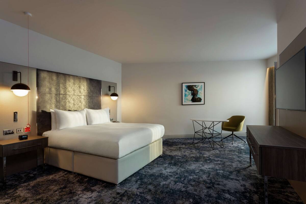 Quarto de hotel moderno com uma cama grande, mesas de cabeceira, escrivaninha, uma cadeira e uma televisão.
