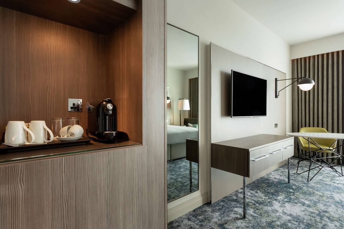 Quarto de hotel moderno com parede em madeira, um espelho grande, televisão de tela plana, mesa com cadeira e um espaço para café.