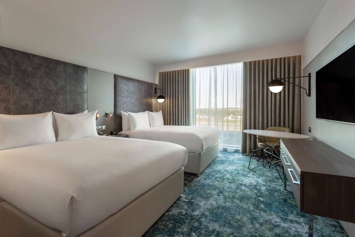 Quarto de hotel com duas camas, uma escrivaninha, televisão e uma janela grande com cortinas.