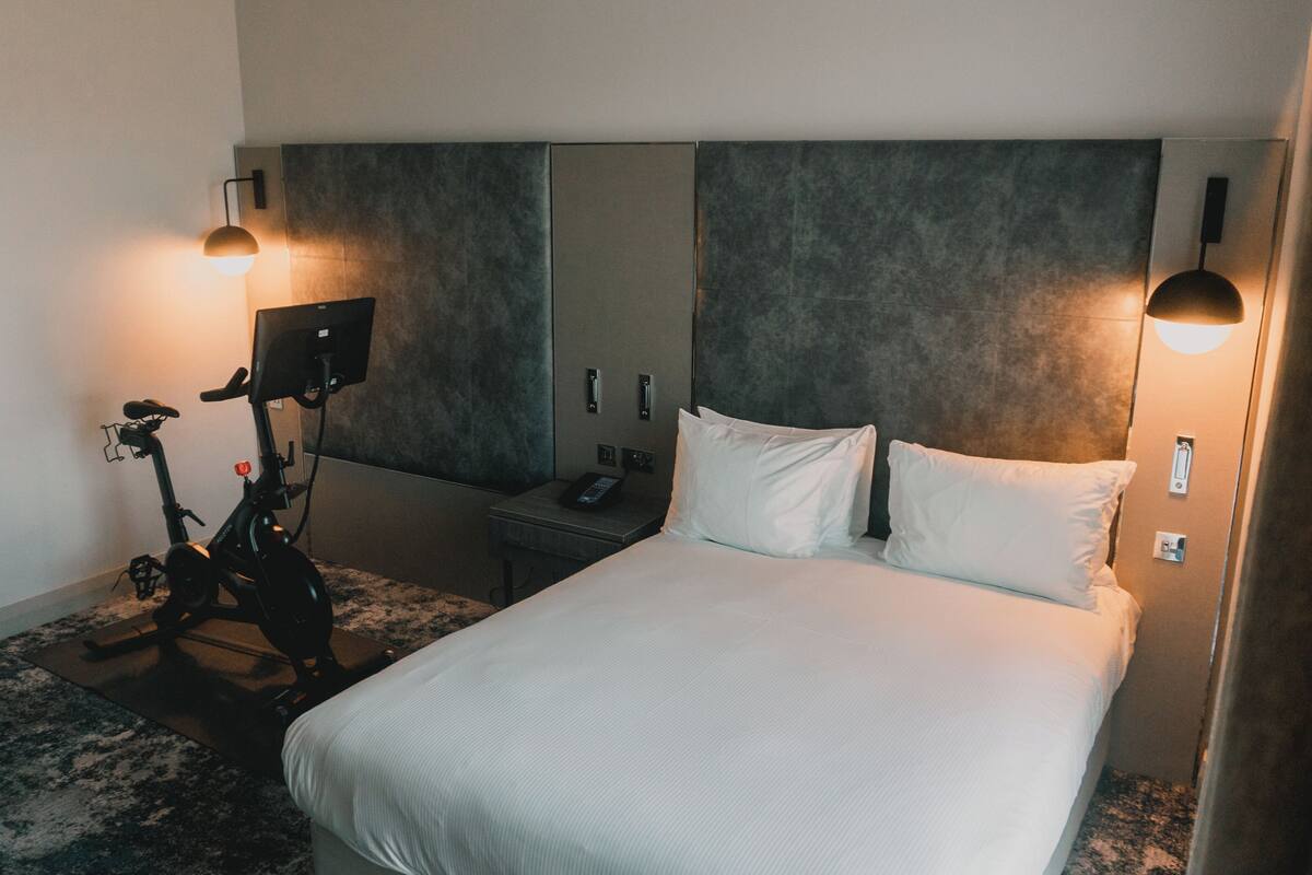Quarto de hotel com uma cama grande, uma mesa de trabalho com telefone, uma bicicleta e duas luminárias fixadas na parede.