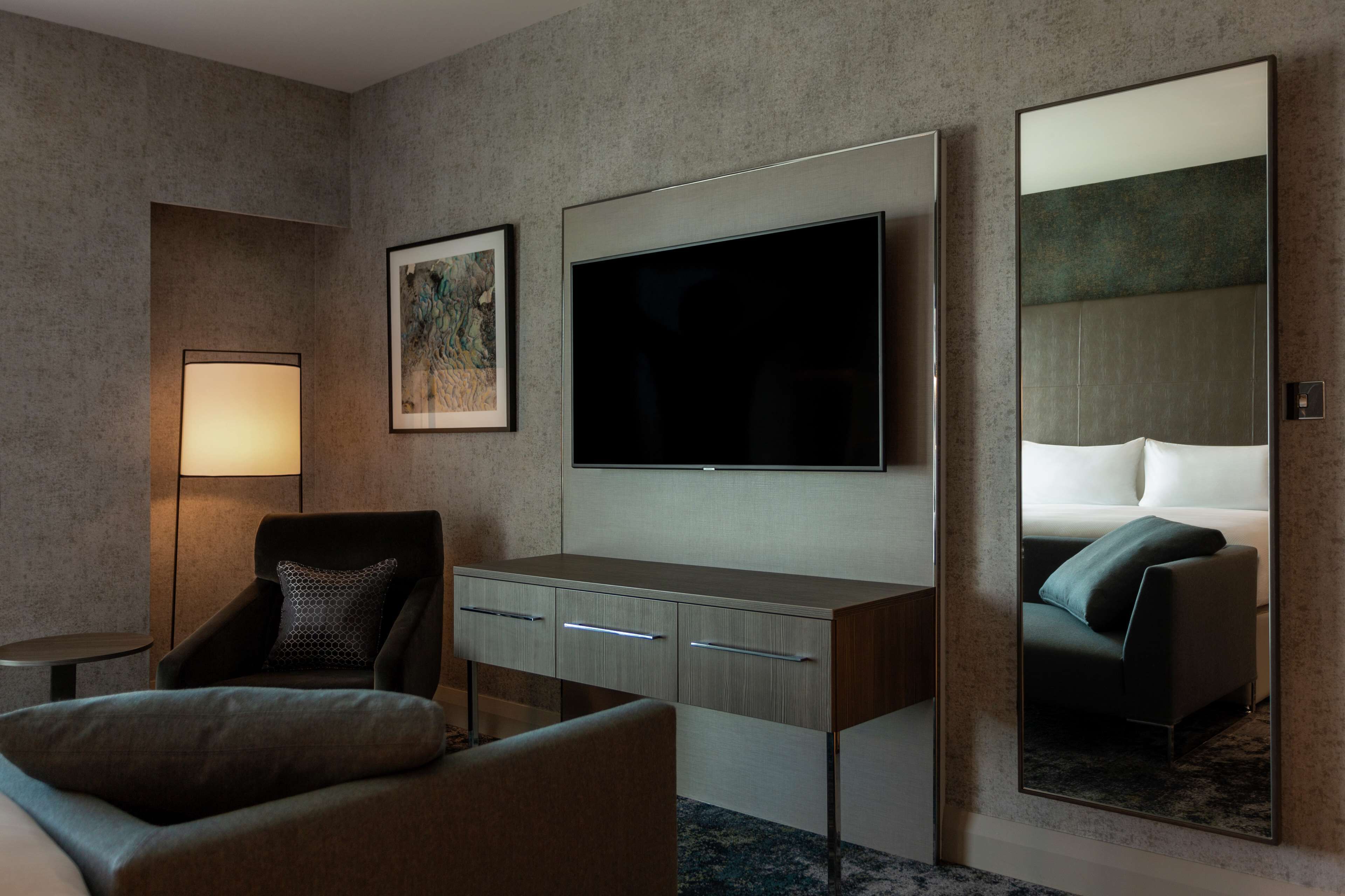 A modern hotel room with a large flat-screen TV mounted on the wall, a comfortable armchair, a small round table, and a large mirror reflecting the room.