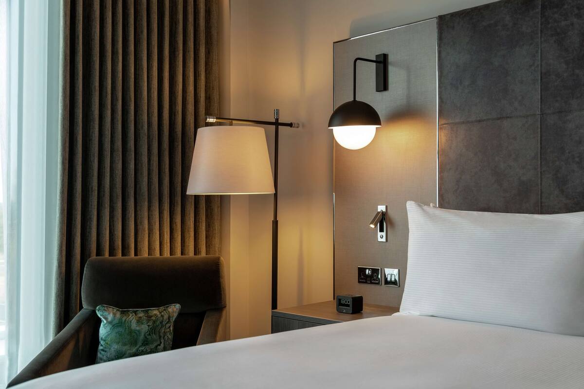 Quarto de hotel com cama, mesa de cabeceira, luminária, cadeira e painel de controle fixado na parede.