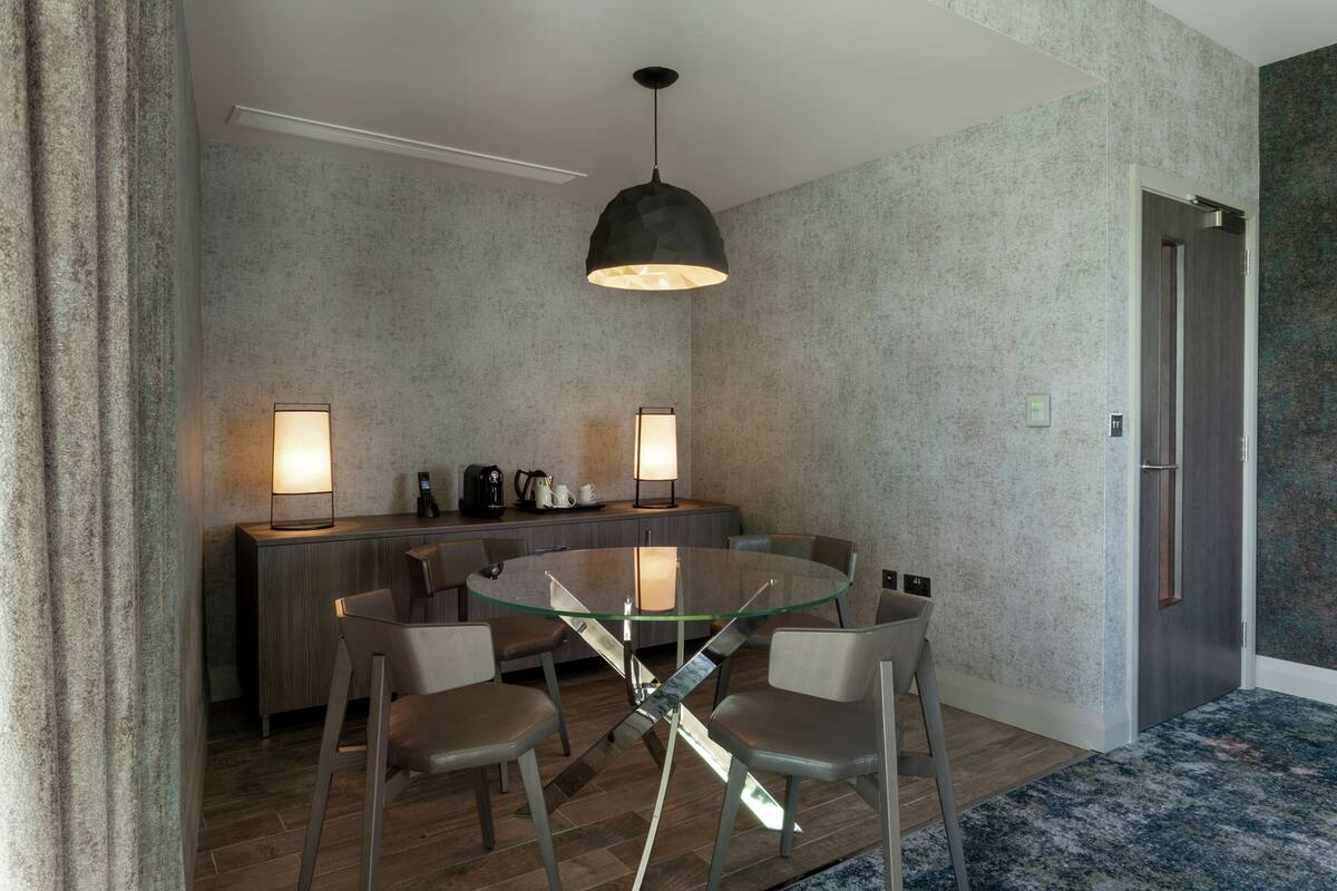 Uma sala de jantar moderna com mesa de vidro, quatro cadeiras, duas luminárias pendentes e bancada de cozinha com eletrodomésticos.