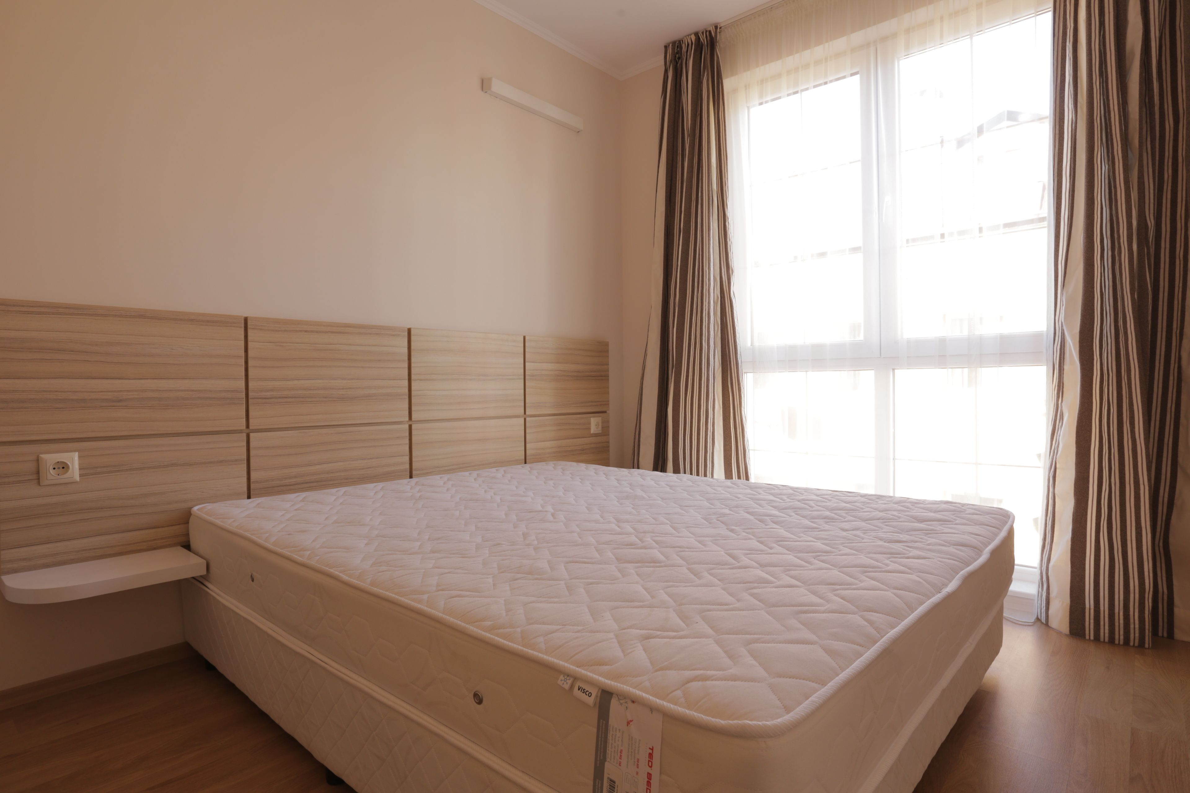 A bedroom with a bed, wooden headboard, a window with curtains, and a bedside table.