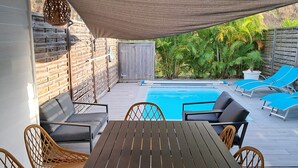 Une terrasse extérieure avec une piscine, une table en bois, des chaises en osier et un espace salon abrité.