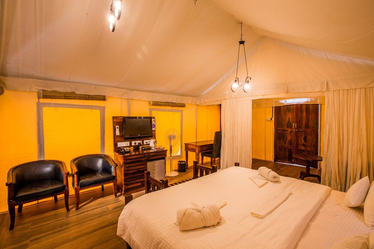 Een tent met een bed, twee stoelen, een bureau met een televisie en een houten garderobe.