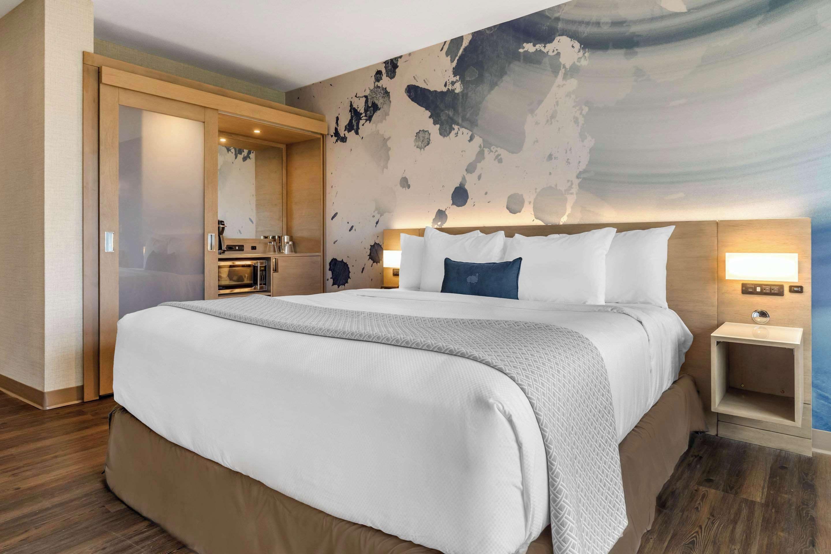 A modern hotel room with a large bed, a nightstand with a lamp, a wooden wardrobe, and a bathroom with a mirror and sink.