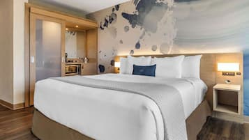 A modern hotel room with a large bed, a nightstand with a lamp, a wooden wardrobe, and a bathroom with a mirror and sink.