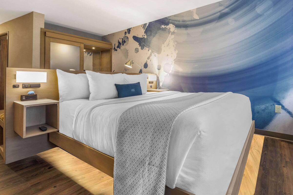 Moderni hotellihuone, jossa on suuri sänky, puinen pääty, yöpöydät lampuineen ja seinällä suuri maailmankartta-aiheinen seinämaalaus.