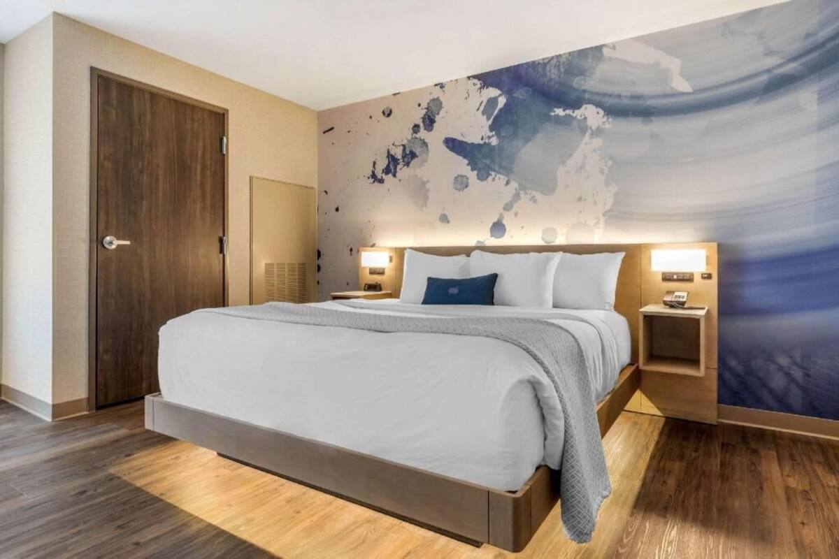 Moderni hotellihuone, jossa on suuri sänky, puulattia, suuri maailmankartta-aiheinen seinämaalaus ja kaksi yöpöytävalaisinta.
