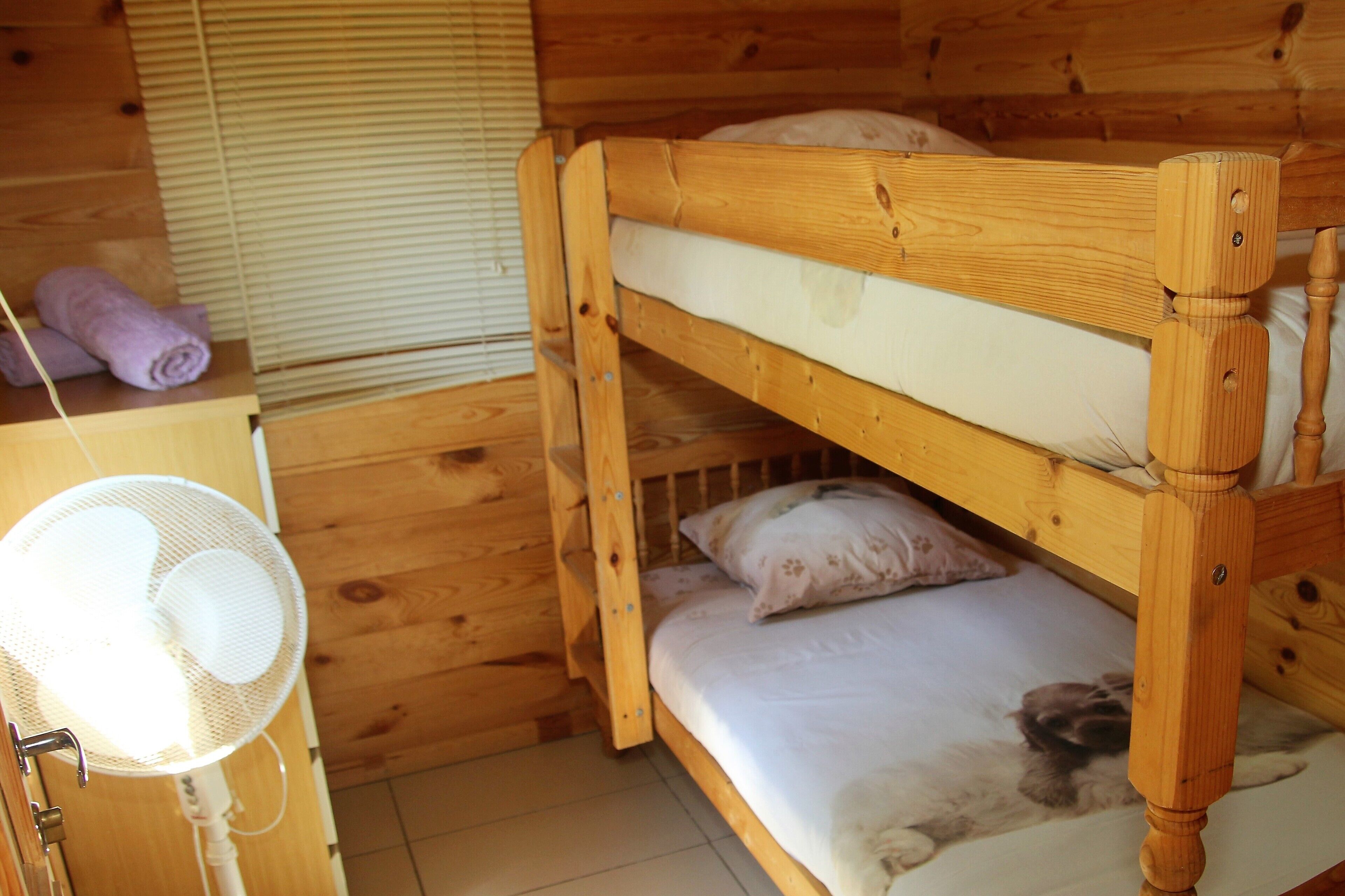 Un lit superposé en bois, avec un ventilateur et des serviettes enroulées posées sur une commode en bois.