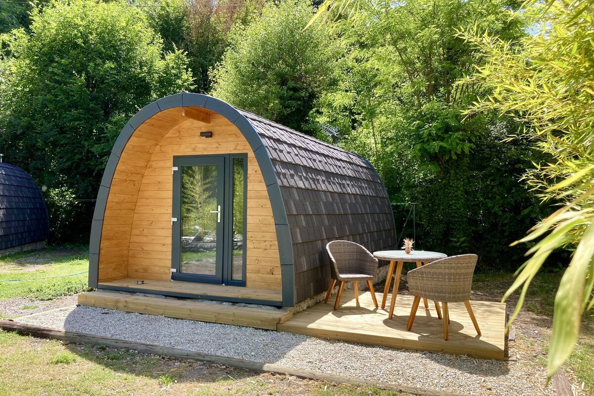 Eine kleine, halbkreisförmige Holzhütte mit Tür und Fenster, neben einem Tisch und zwei Stühlen.