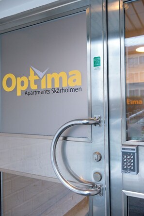 A metallic door with a handle and a keypad lock, featuring the logo and name 'Optima Apartments Skärholmen'. - Optimal Apartments Skärholmen (Skärholmen)