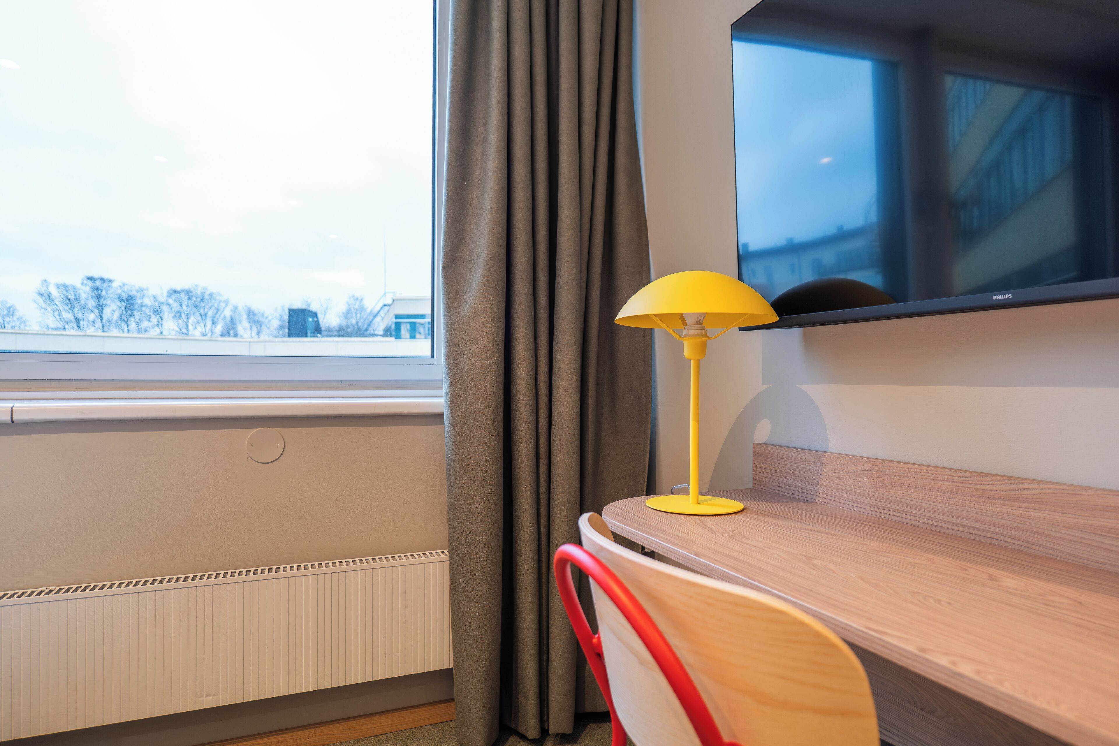 Una habitación con un escritorio de madera, una lámpara amarilla, una silla roja, una ventana con cortinas y vistas a edificios.