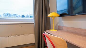 Una habitación con un escritorio de madera, una lámpara amarilla, una silla roja, una ventana con cortinas y vistas a edificios.